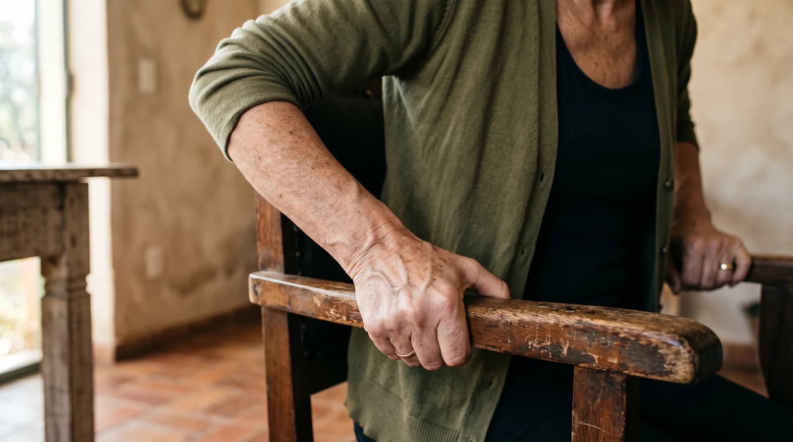 Mãos de uma mulher idosa apoiadas no braço de uma cadeira de madeira para se levantar